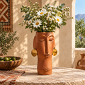 Terracotta face vase with gold disc earrings filled with daisies in a sunlit Mediterranean courtyard with tribal rug wall hanging